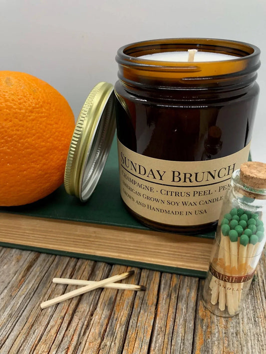 Soy wax candle in amber jar labeled Sunday Brunch with an orange, matches, and book on a rustic table
