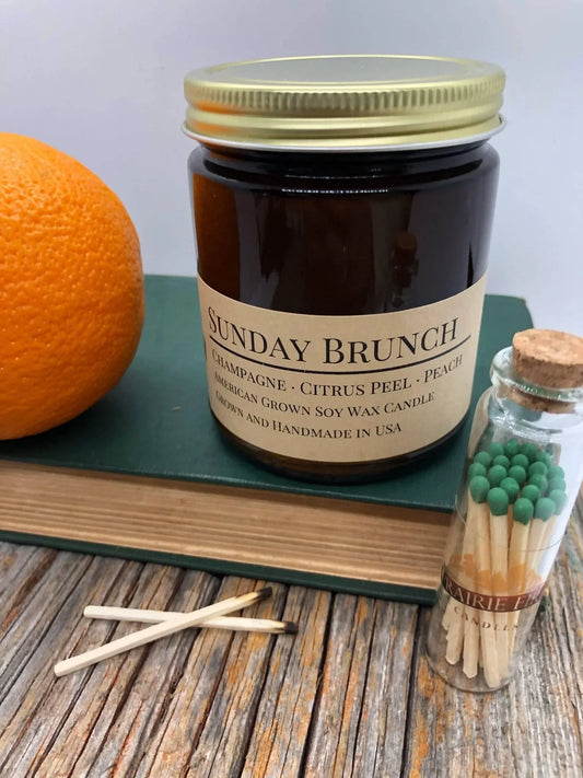 Sunday Brunch soy wax candle in amber jar with orange, book, and green-tipped matches