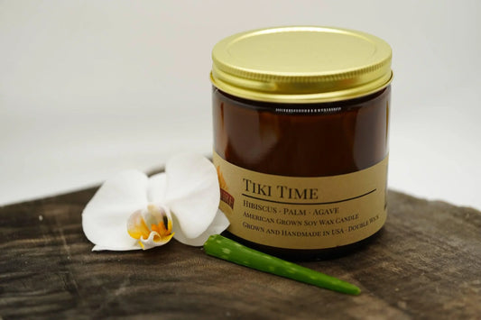 Tiki Time soy wax candle in amber jar with gold lid, orchid flower and green leaf on wood surface.