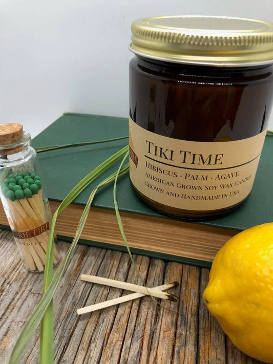 Tiki Time soy wax candle in amber jar with matches, book, lemon, and grass on wood table