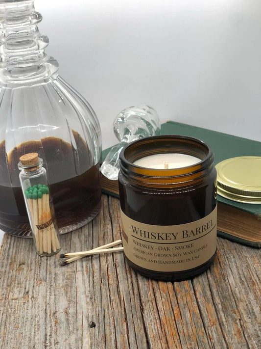 Whiskey Barrel soy wax candle in amber jar beside glass decanter, matches, and wood surface