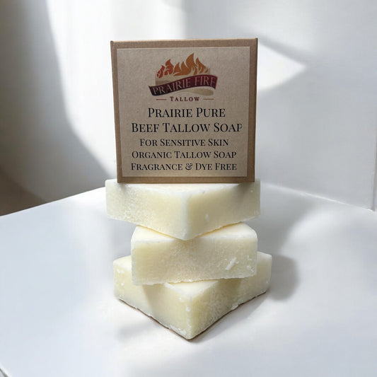 Three natural beef tallow soap bars stacked on a white surface in sunlight, with packaging labeled for sensitive skin, organic, fragrance and dye free.